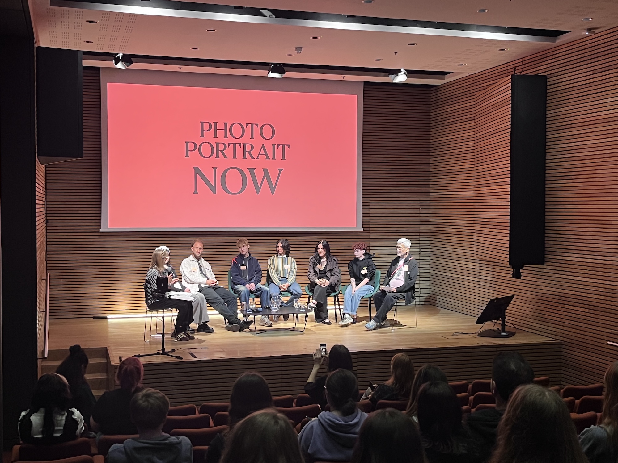 A panel of seven people seated on stage in front of a large screen reading 'PHOTO PORTRAIT NOW', with an audience in the foreground.