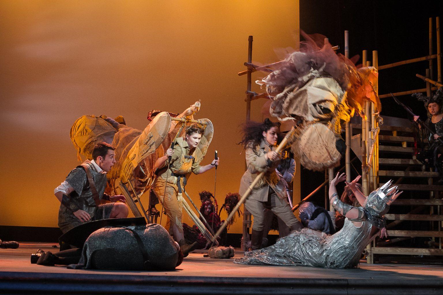 Actors on stage, two serving as the head and body of a lion. An actress in costume as an ice queen is on the ground and cowering before the lion's head.
