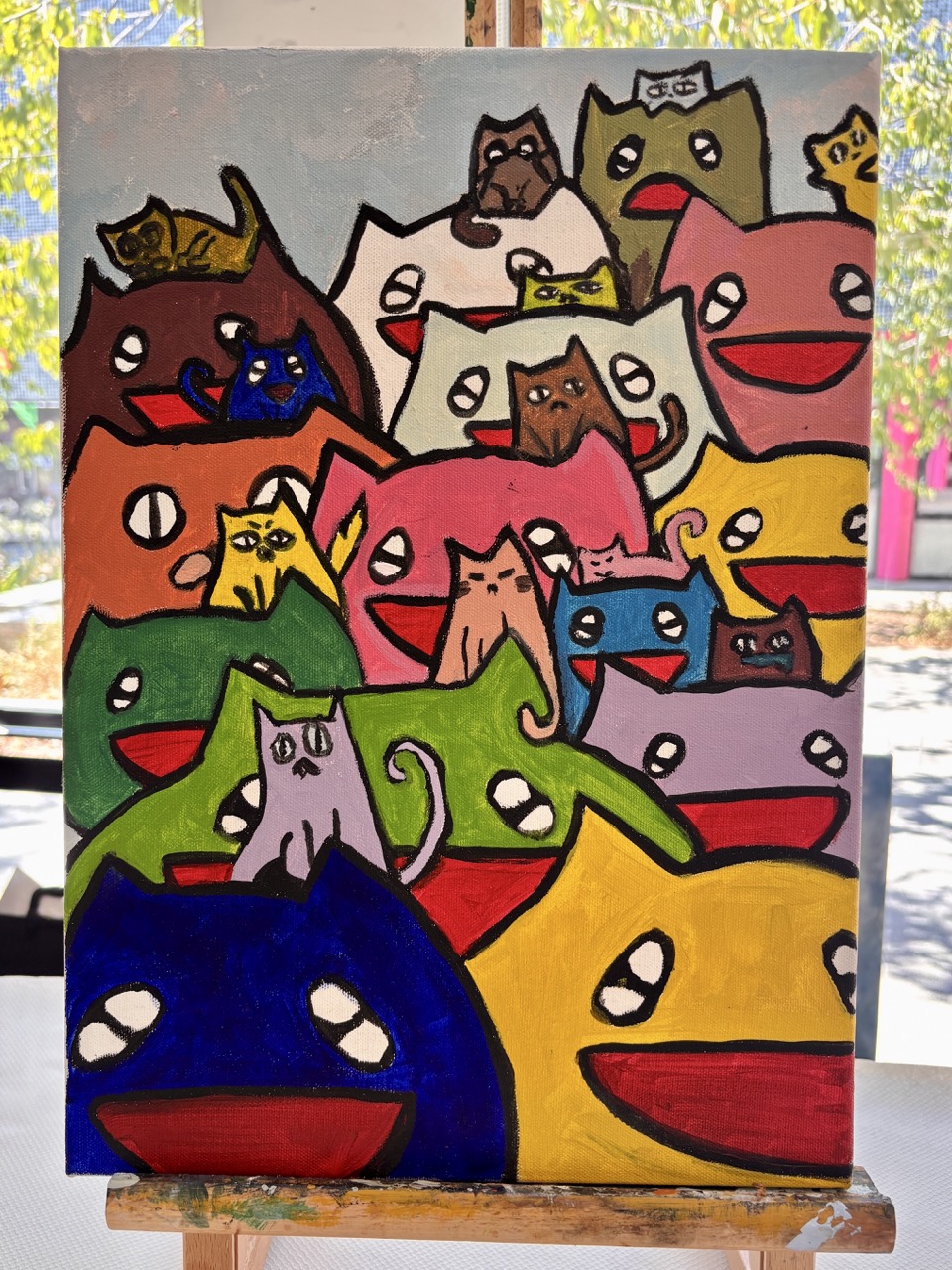 A colourful painting of overlapping cartoon cats with wide open mouths on a canvas, displayed on an easel.