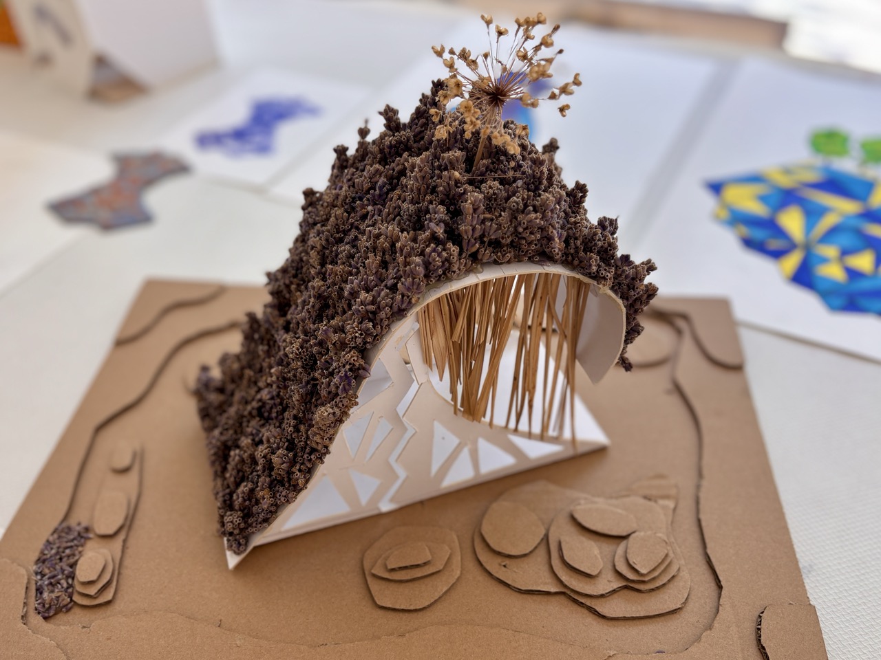 A textured miniature arch sculpture covered in dark material and supported by wooden sticks, displayed on a brown base with layered cut-out shapes.