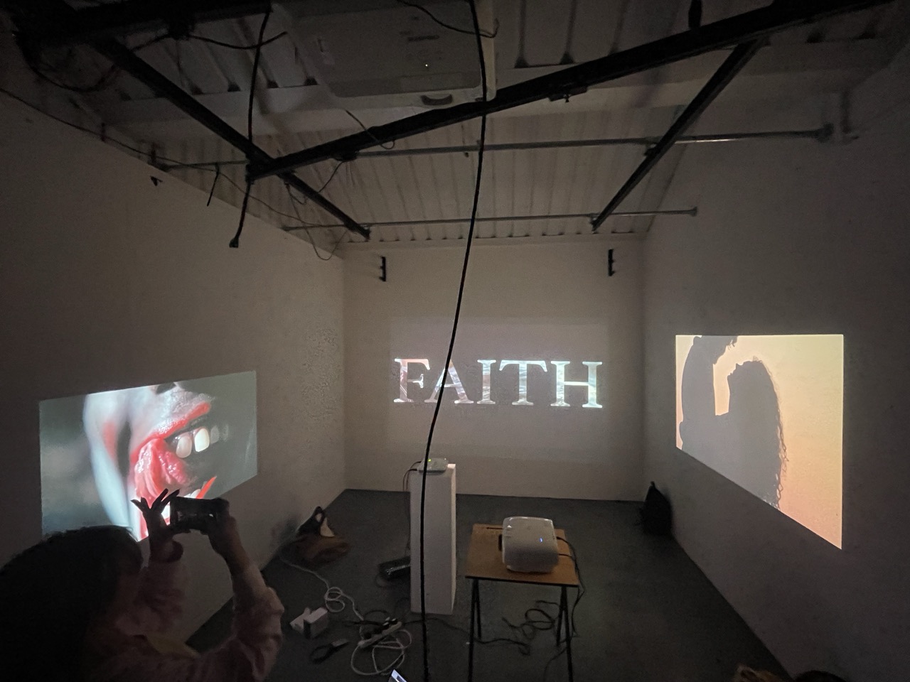 An art installation in a dim room with the word FAITH projected on the central wall, alongside two side projections and a small table with equipment.