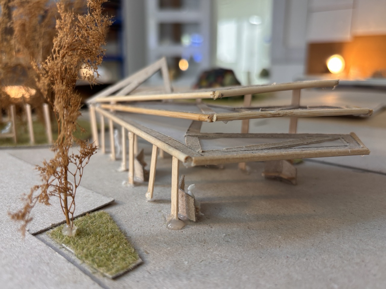 A small architectural scale model of a raised pavilion with a slatted roof, set on a grey base beside miniature trees and grass patches.