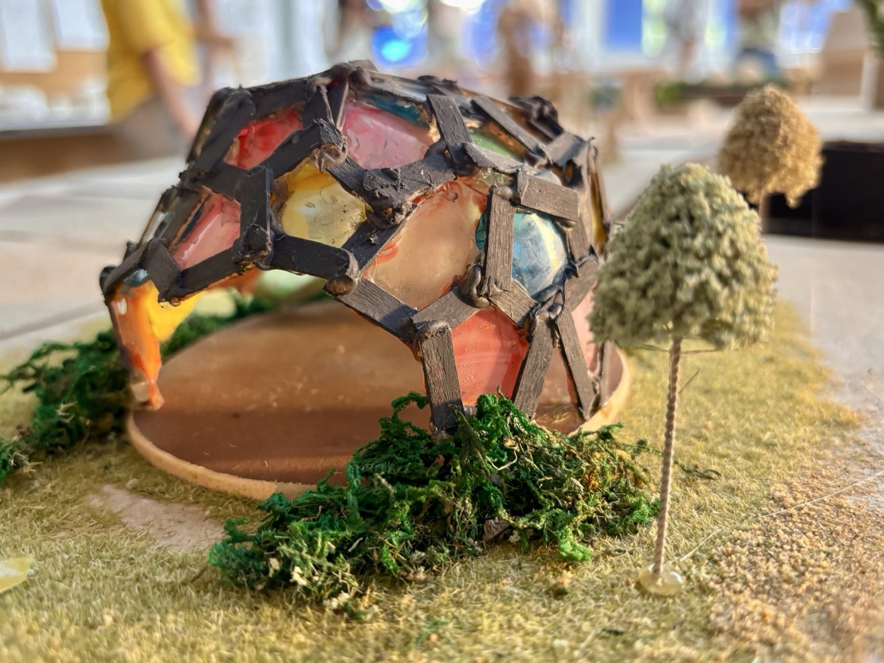 A handmade miniature dome structure with stained glass-like panels sits on a model landscape with moss and small trees.