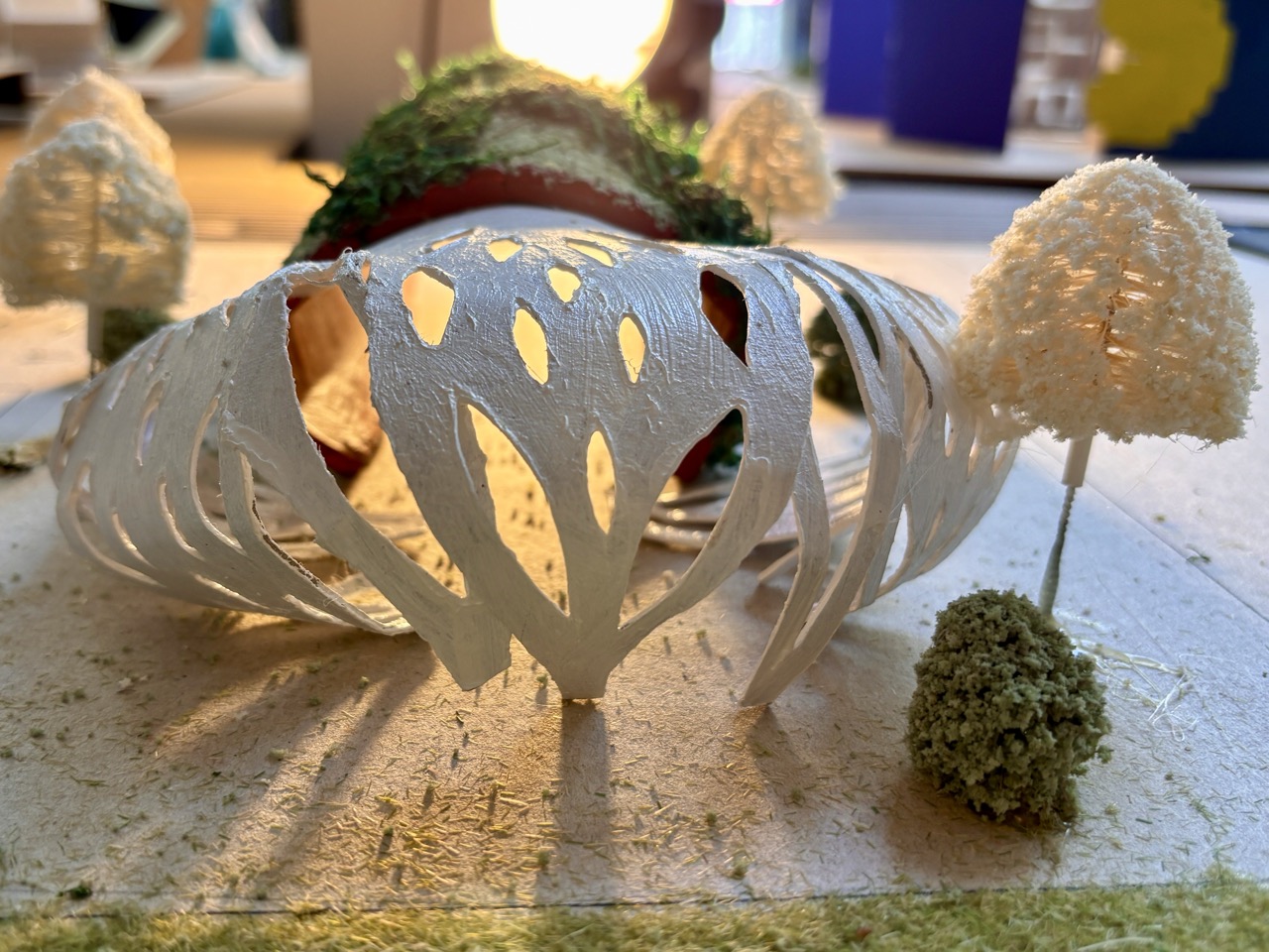 A small white architectural model with curved cut-out panels sits on a tabletop, surrounded by miniature tree shapes and warm backlighting.