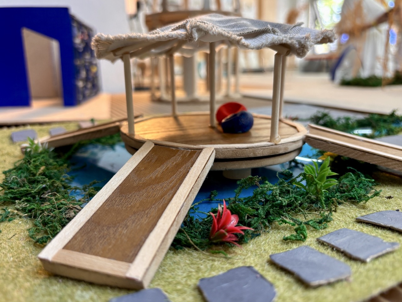 A small wooden model pavilion with a ramp and cloth roof sits on a landscaped display with miniature grass, stepping stones and a tiny red flower.