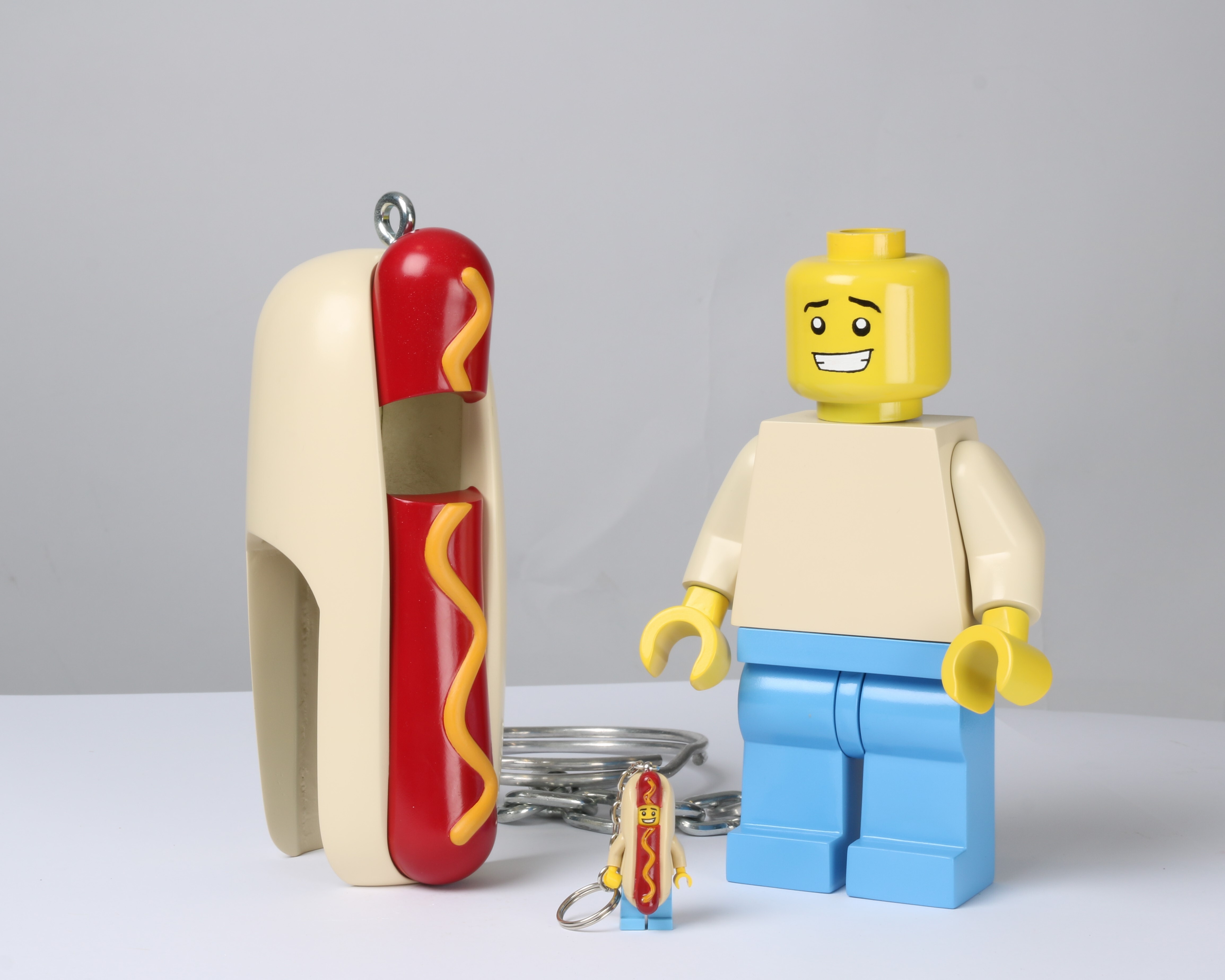 Giant LEGO minifigure with yellow head and white top next to oversized hot dog in bun with red sausage and yellow mustard squiggles. A miniature LEGO minifigure keyring wearing a hot dog costume sits between the two larger models.