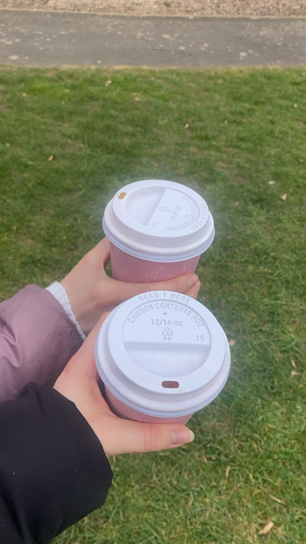 Hands holding two pink takeaway coffee cups with white lids on green grass with grey pavement in background.