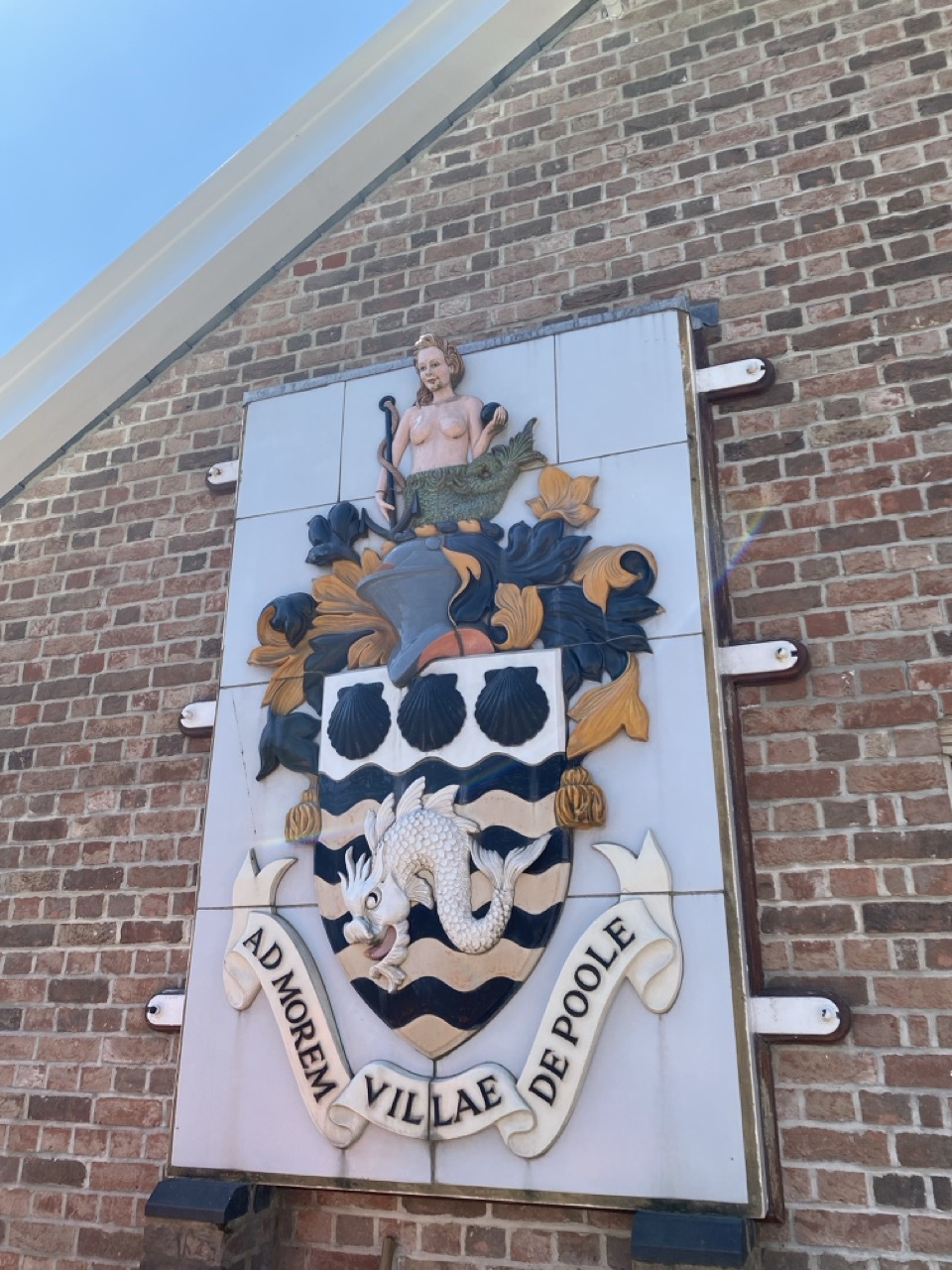 Poole Museum coat of arms logo on side of a brick wall, with waves and a mermaid on top.