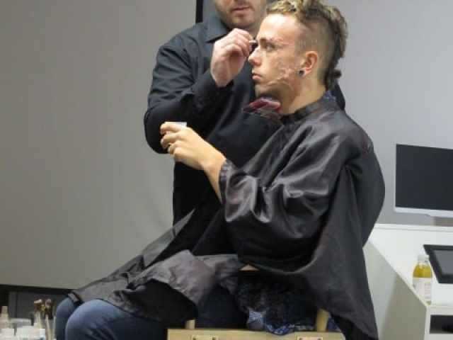 A make-up designer applies make-up to a model with prosthetic facial scars sitting in a chair.
