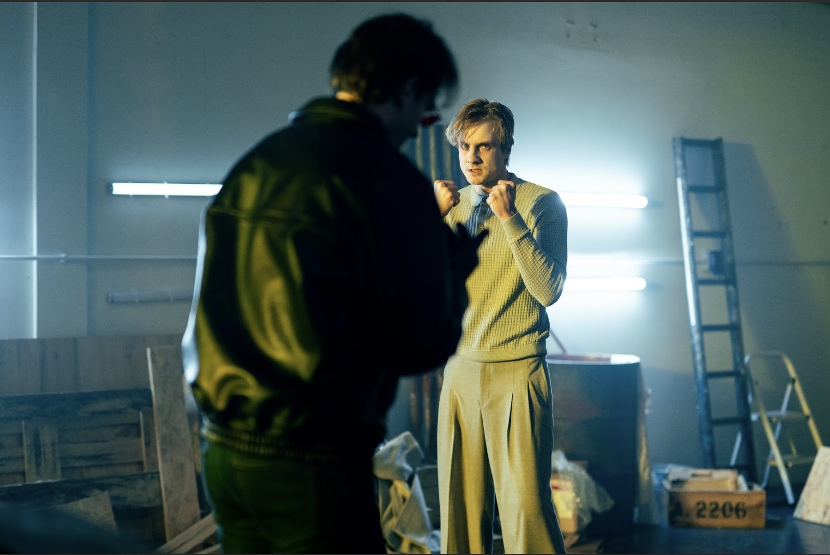 A man in a beige knitted top and trousers stands with raised fists in an industrial room, facing another man in the foreground, with fluorescent tube lights and a ladder in the background.