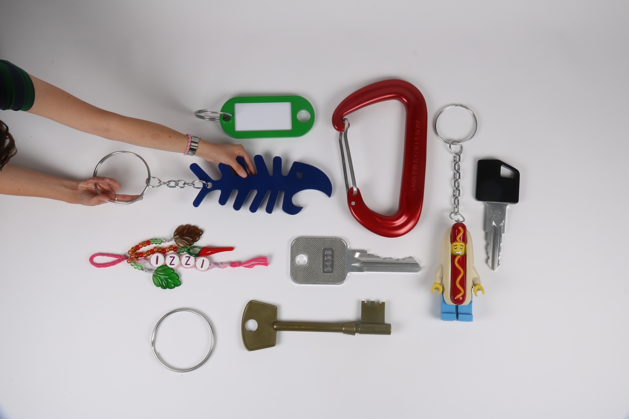 Various keychains and model keys scattered on white surface: hands holding green key tag, blue fish-shaped item, red carabiner, keys, whistle, and colourful accessories.