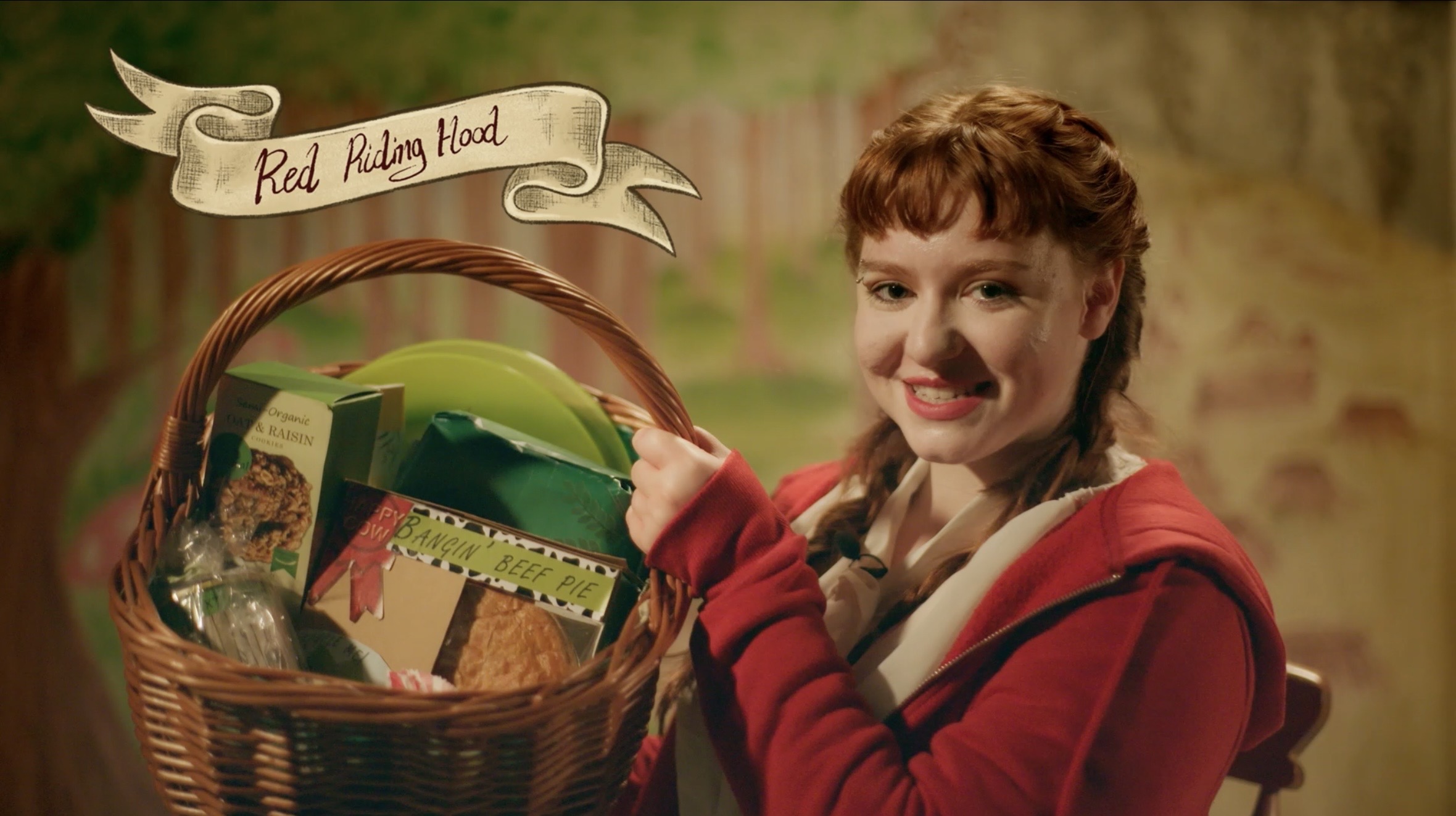 A woman in a red hooded cloak holds a wicker basket of groceries beside a banner reading Red Riding Hood, against a rustic backdrop.