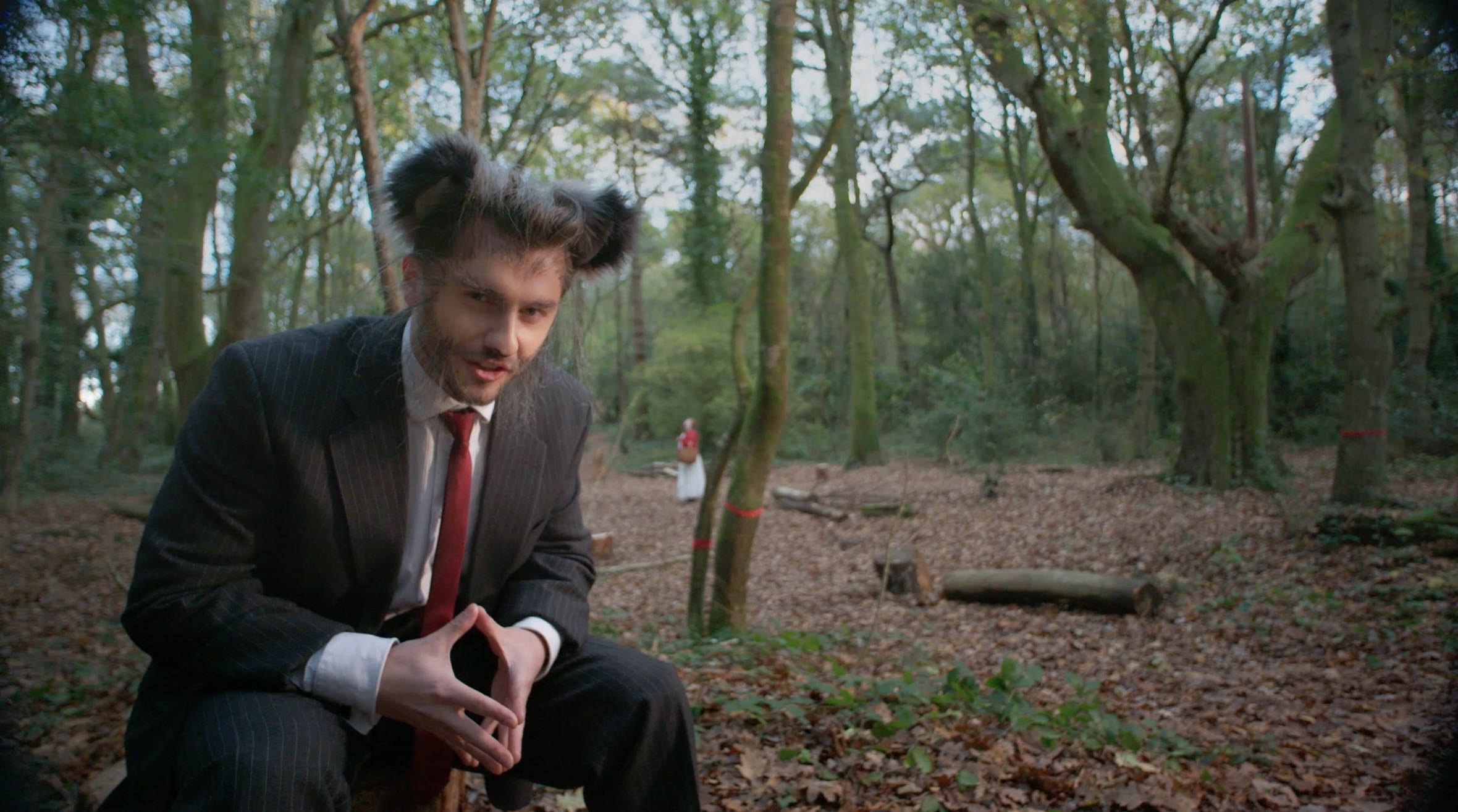 A man in a dark suit and red tie sits on a log in a wooded area, with trees, leaf litter and a distant figure in the background.