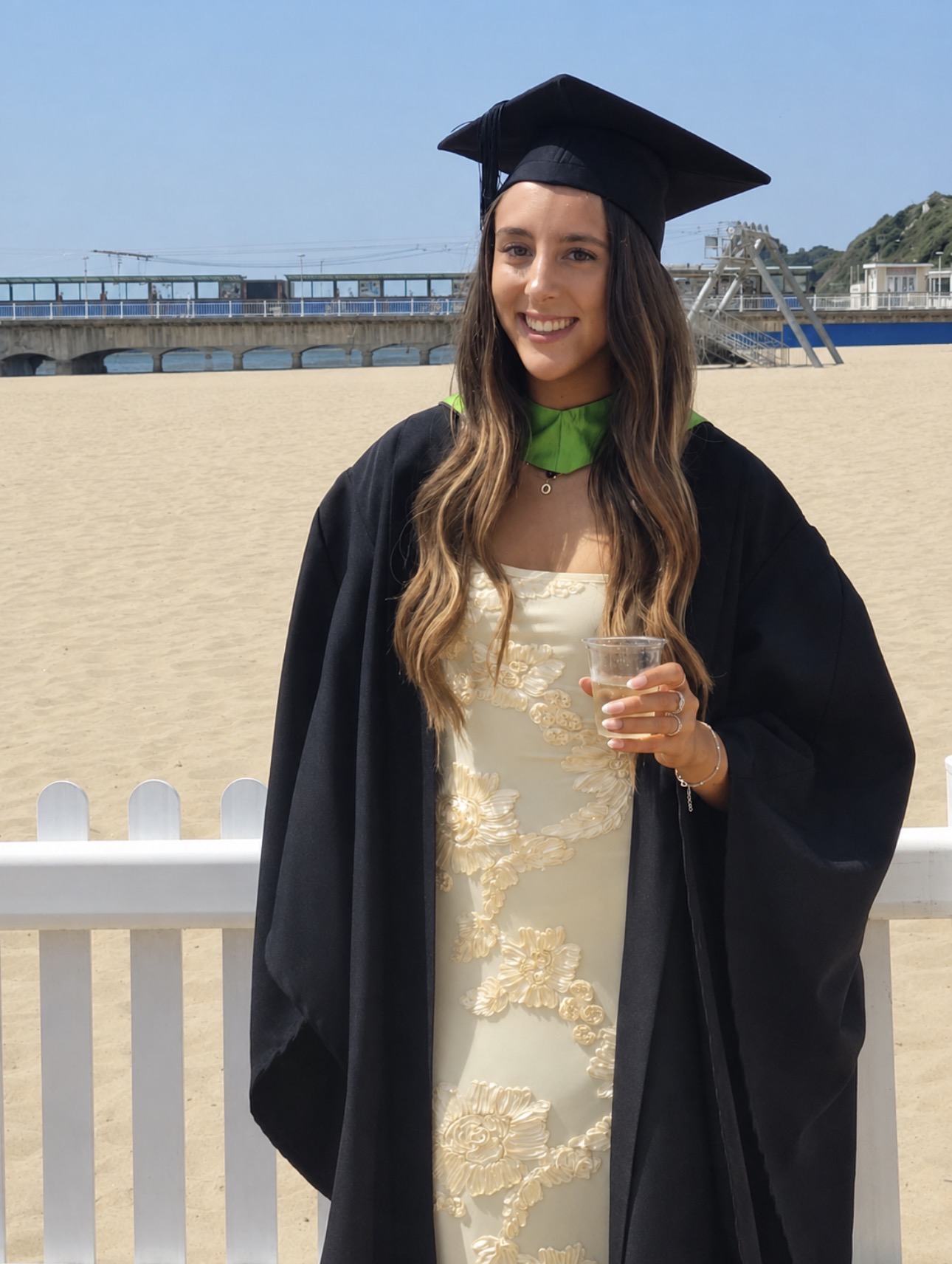 A woman in a graduation cap and gown stands by a beachside railing, holding a drink with a bridge and sand in the background.