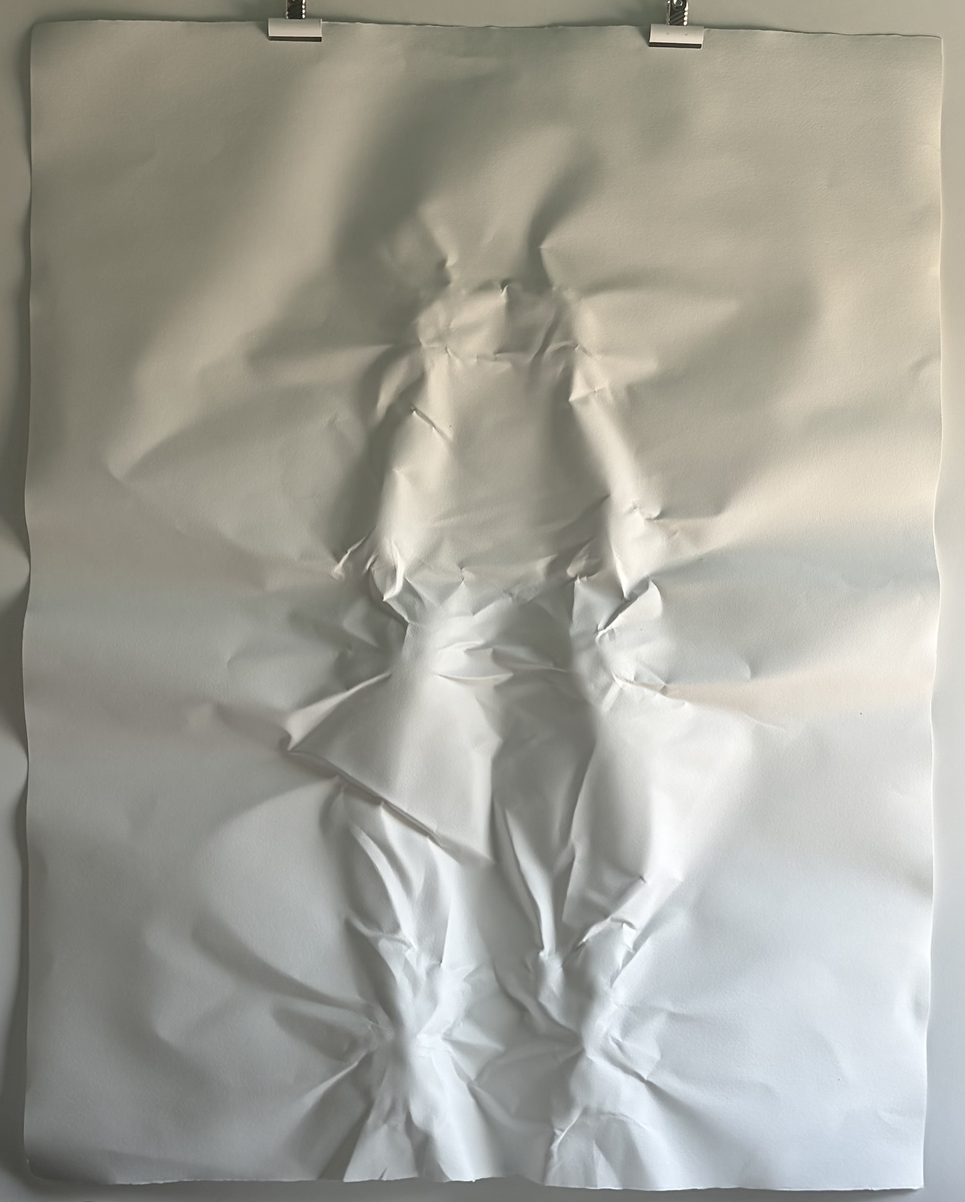 A sheet of silver foil or paper clipped at the top, with crumpled folds forming a human-like silhouette against a plain background.