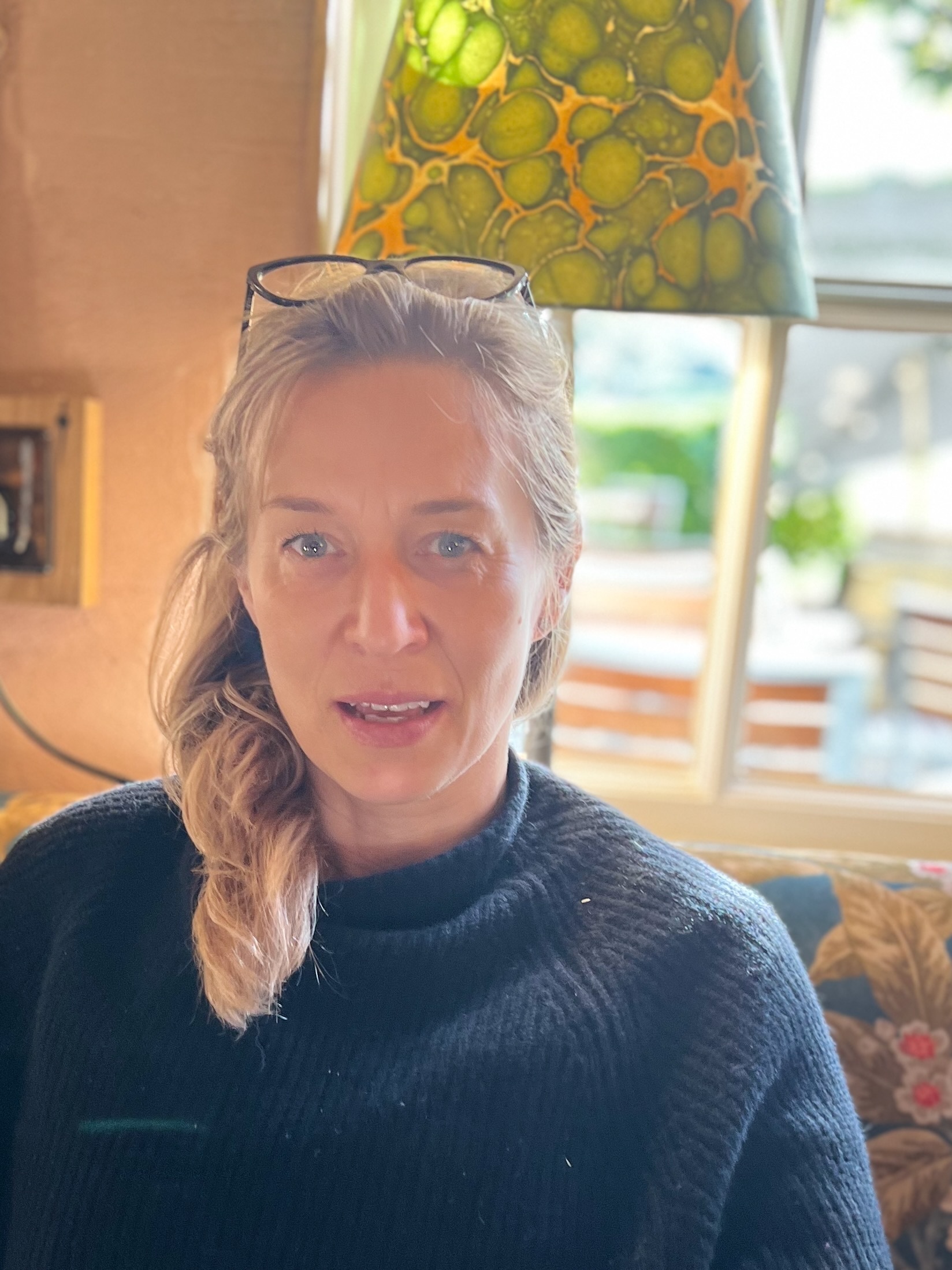 Woman with blonde hair and glasses on head wearing dark jumper, sitting in bright room with green and yellow lampshade above.