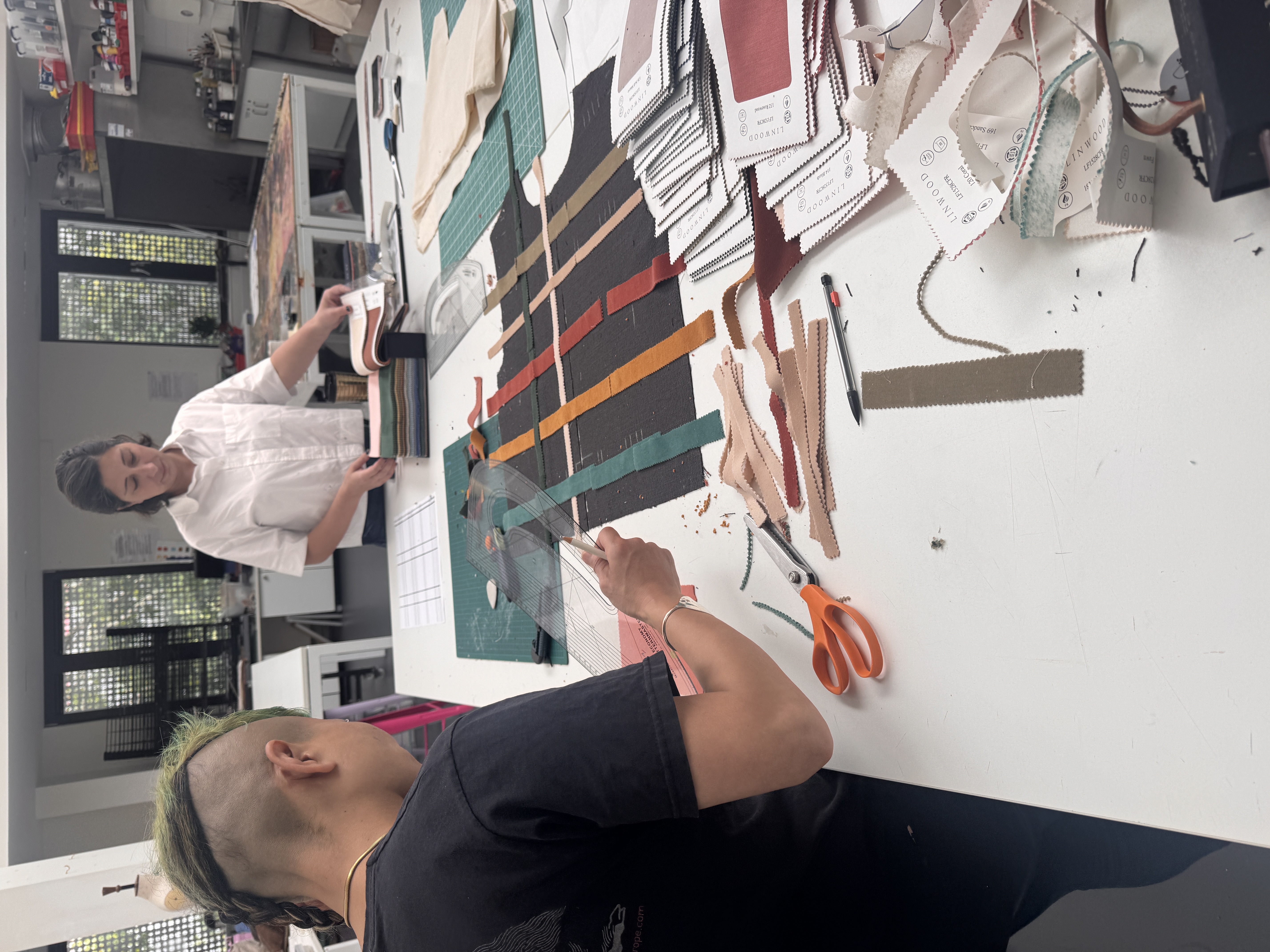 Two people work at a large table covered with paper pattern pieces, fabric strips and scissors in a design studio.