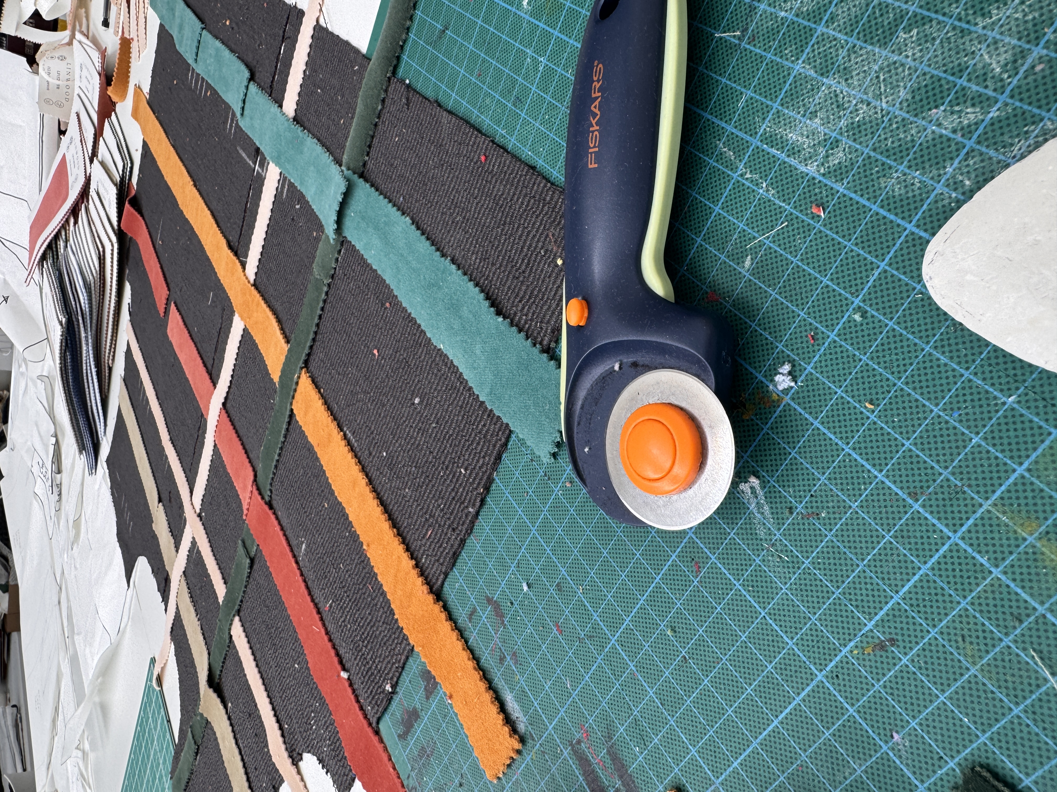 A rotary cutter rests on a green cutting mat beside strips of black fabric with coloured woven lines, with white pattern pieces partly visible at the edge.
