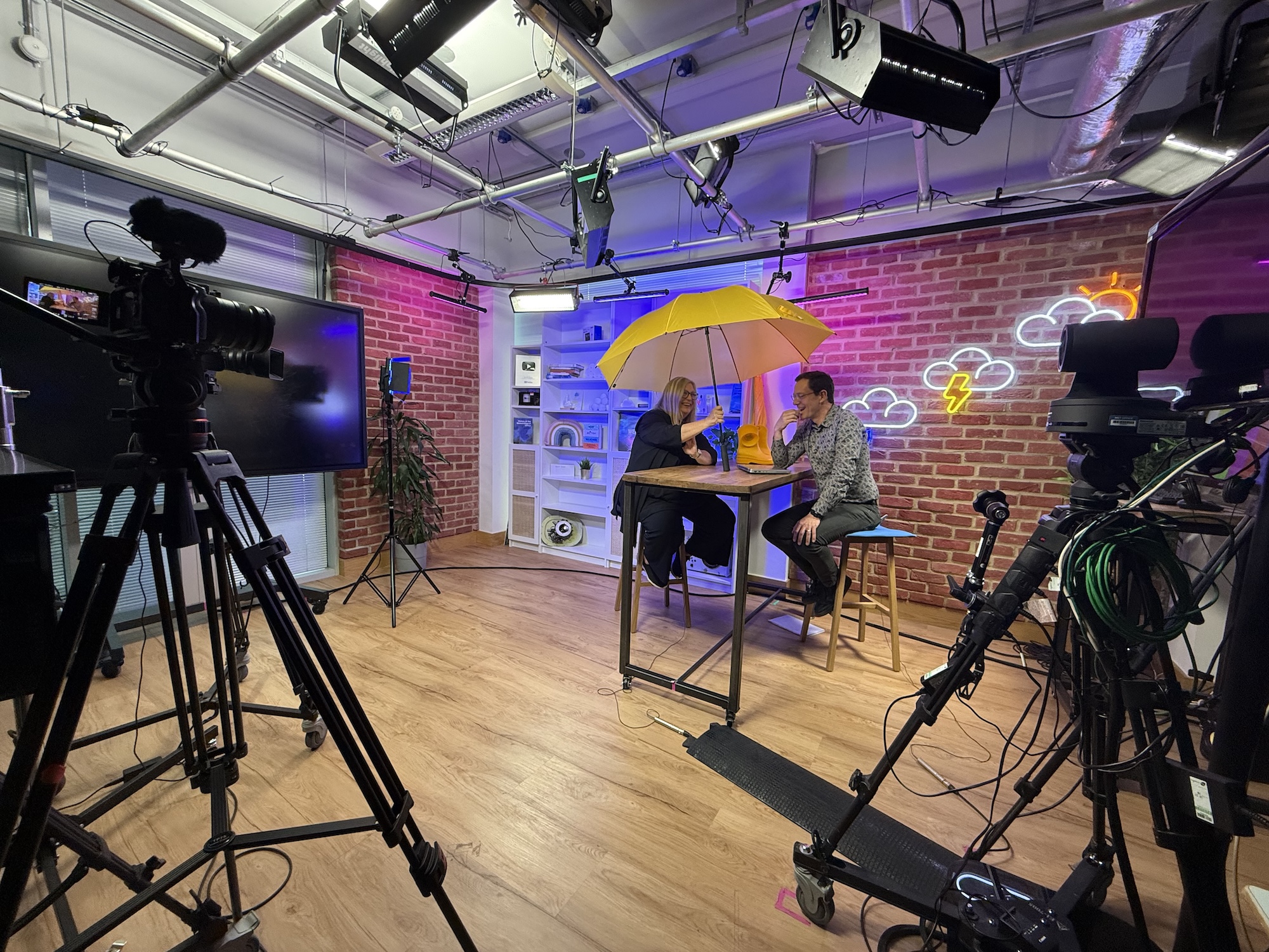 Two people sit at a high table under a yellow umbrella in a studio, framed by cameras on tripods and neon cloud and lightning signs on a brick wall.