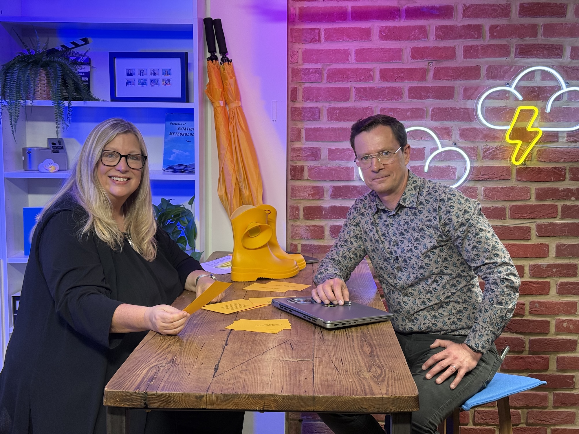 A. Stevens and A. Deakin sit at a wooden table with papers and a laptop, in a studio with shelves, a brick wall and neon cloud and lightning signs.