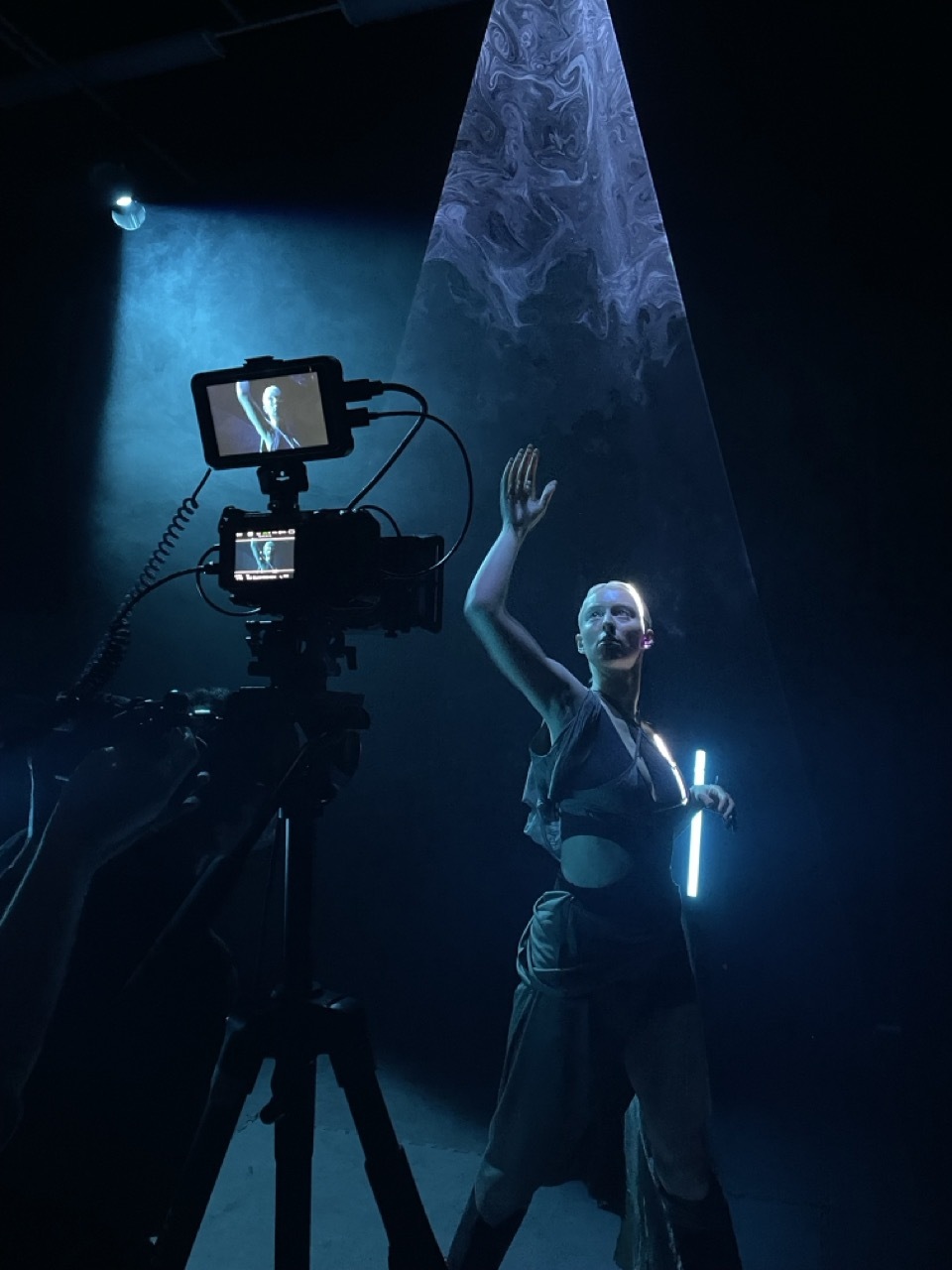 Film set with blue lighting, camera on tripod, person with raised arm holding light sabre, draped fabric backdrop visible above.