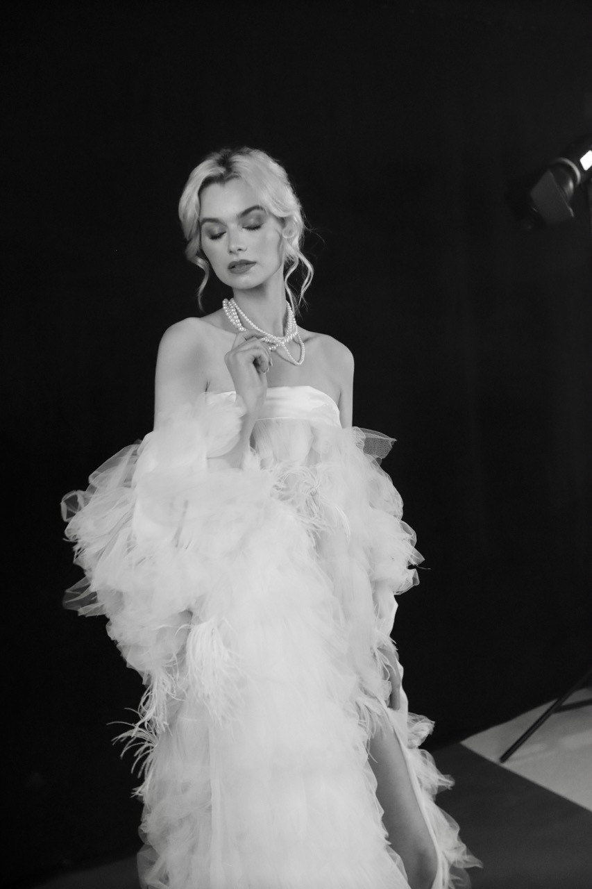 Black and white image of woman in voluminous white ruffled gown with statement necklace, blonde hair in updo, against dark background.
