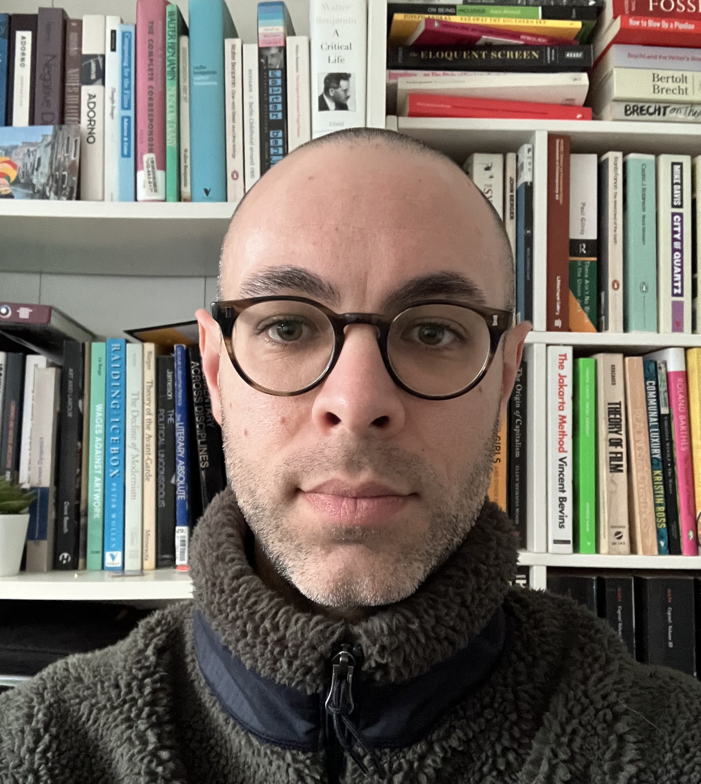 Bald man with round glasses and grey fleece jacket sits in front of white bookshelves filled with multicoloured books.