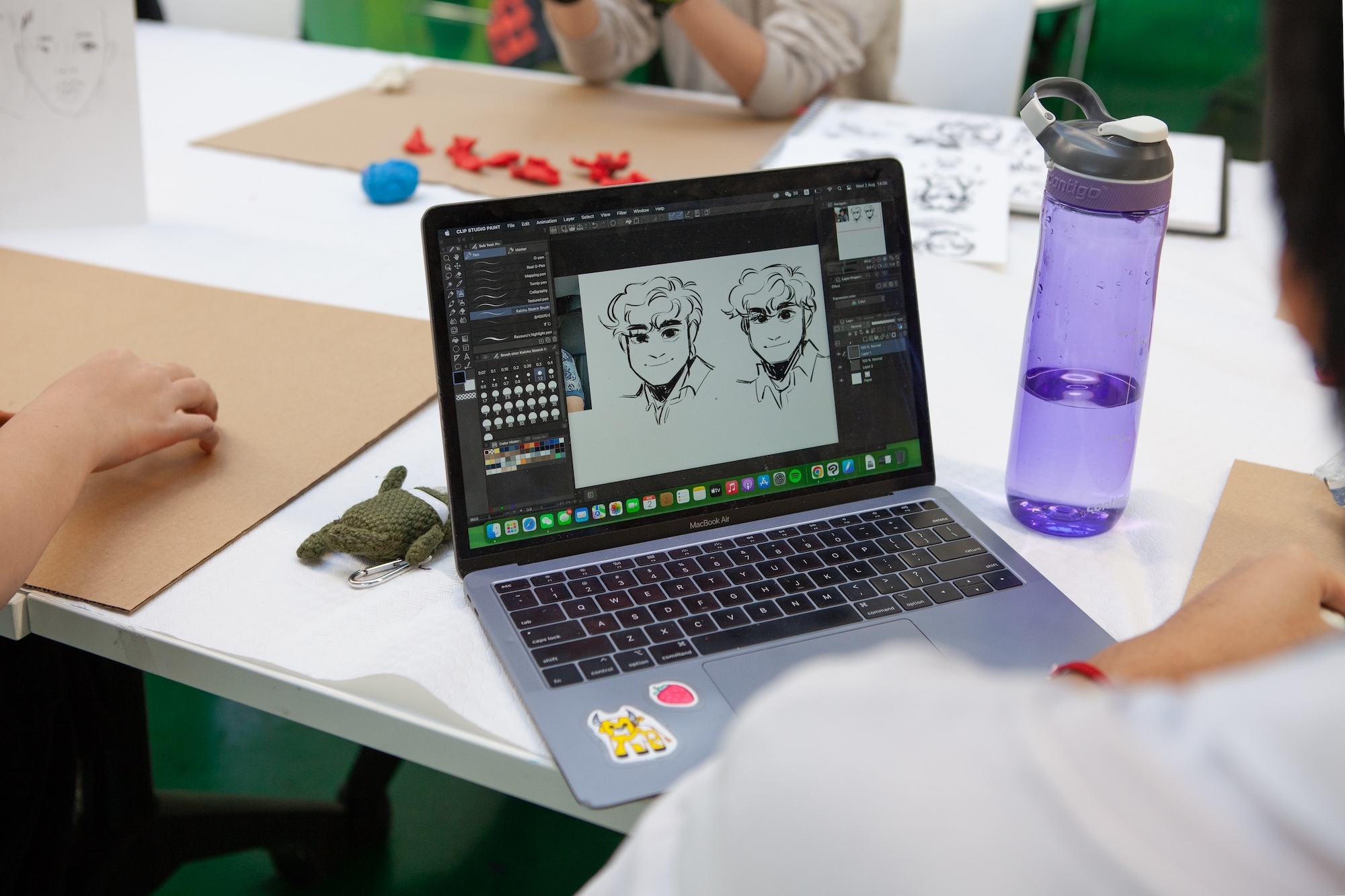 MacBook displaying digital art software with two character sketches on screen, purple water bottle beside it on white desk.