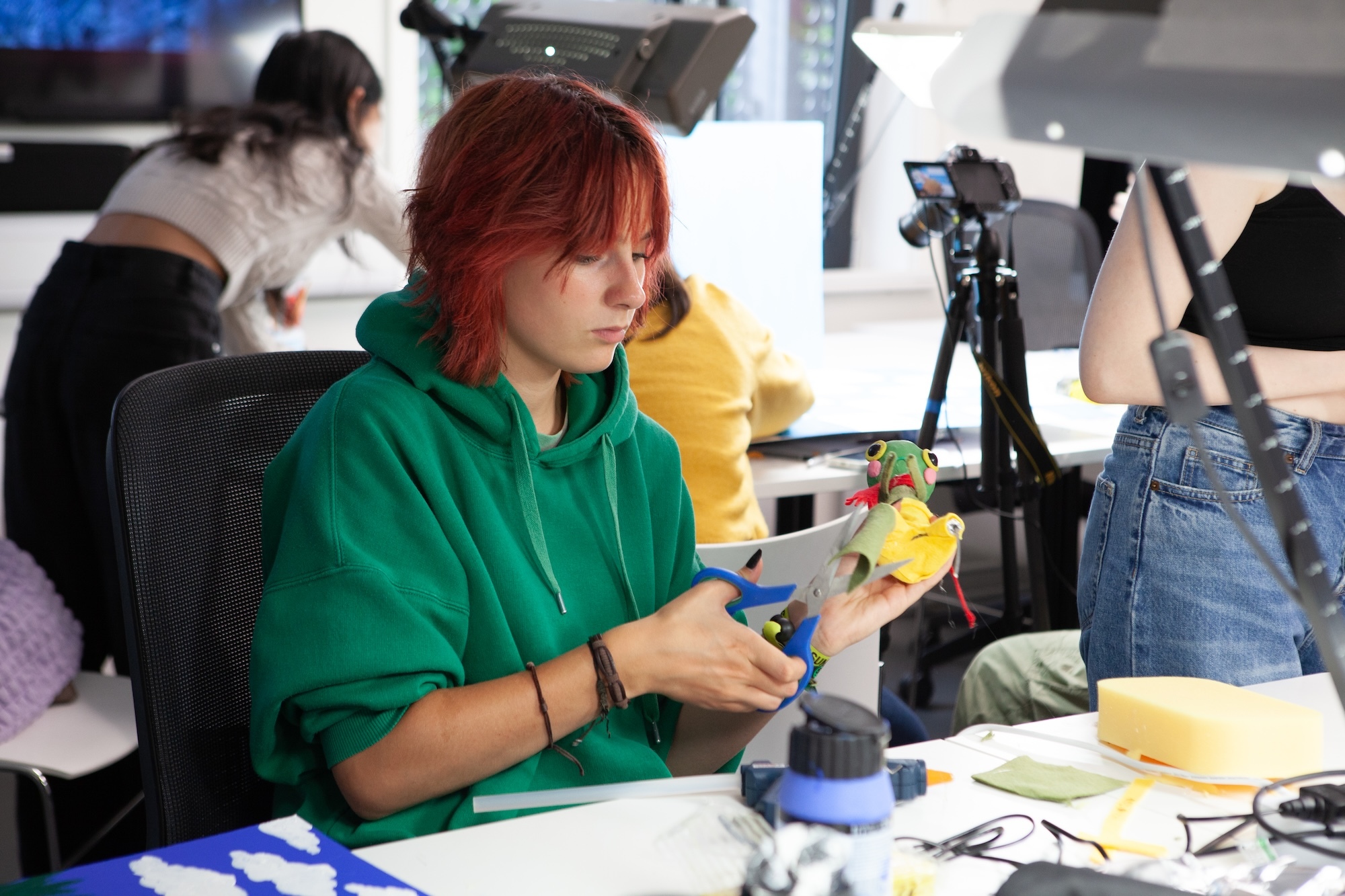 Person in green hoodie with red hair working at desk with camera equipment, papers, and various materials scattered around workspace.