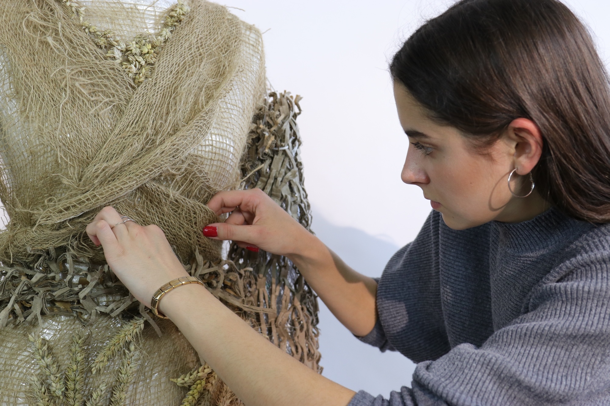 Woman in grey jumper adjusting olive green textured fabric with fringe on dress form against white background.