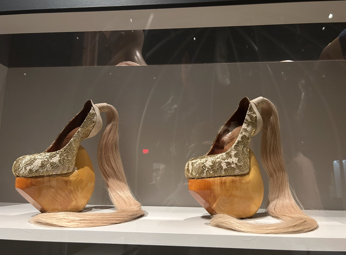 Two avant-garde platform shoes in museum display case with curved wooden heels, metallic uppers, and orange wedge sections.