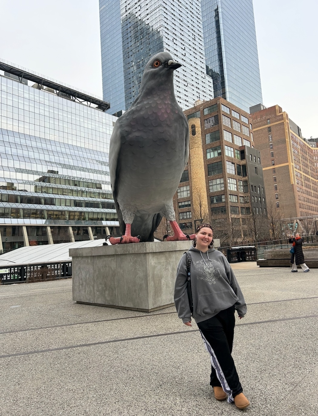 Woman posing beside large grey pigeon sculpture on concrete plinth in urban plaza with glass and brick buildings in background.