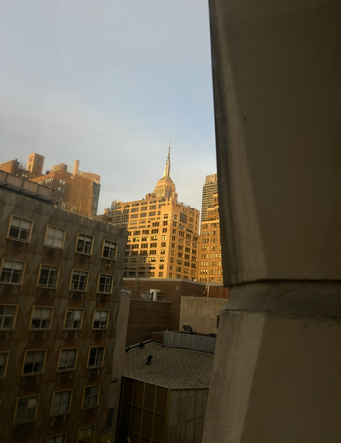 View from window showing urban buildings bathed in golden sunlight, including Empire State Building in distance, with grey building frame visible on right.