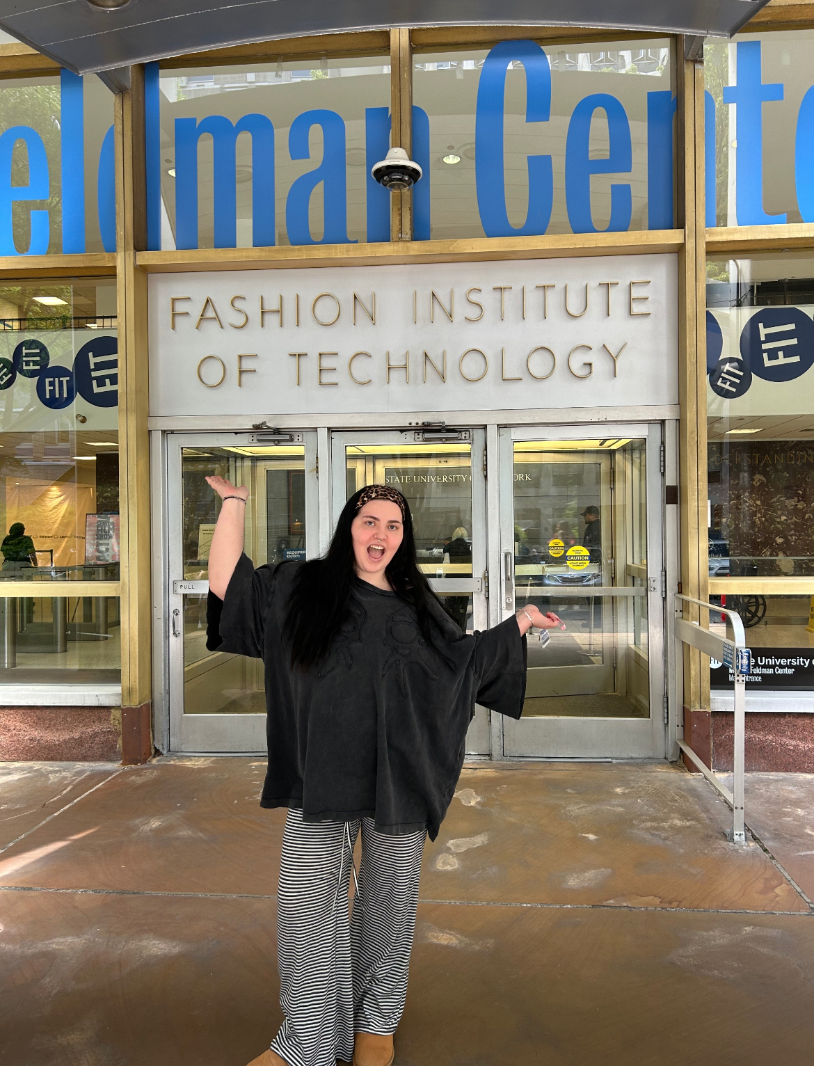 M Koch celebrating with raised arms outside Fashion Institute of Technology building entrance.