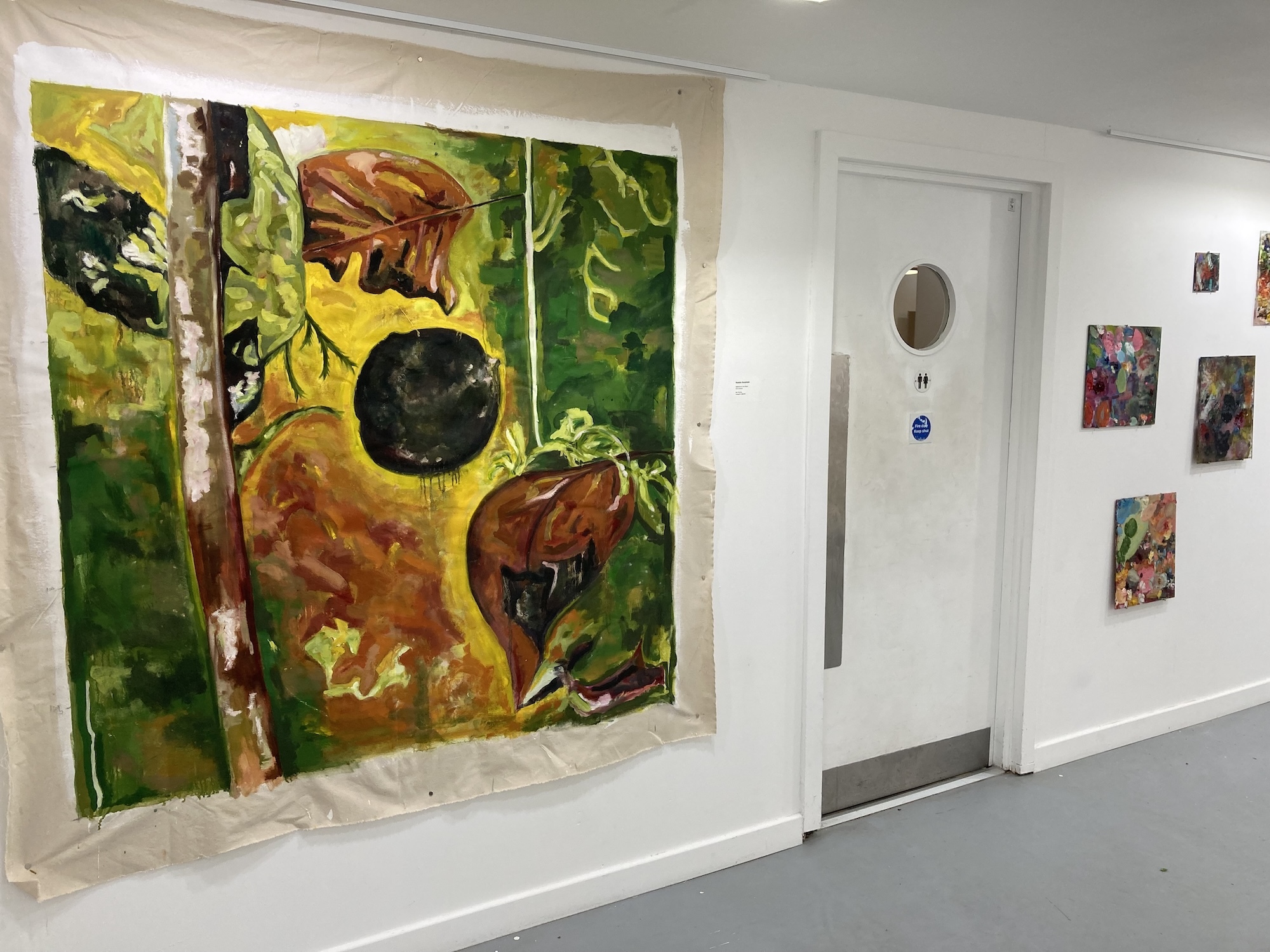 Gallery wall displaying large colourful painting with yellow, green, brown and black abstract shapes alongside smaller framed artworks.