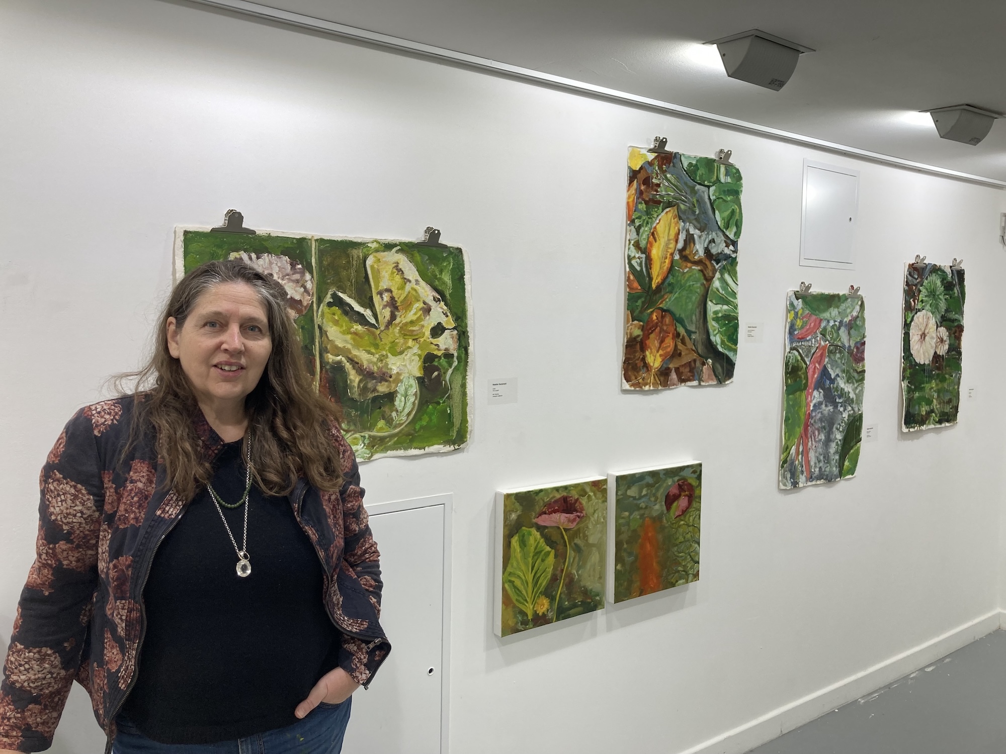 Woman with long dark hair in patterned cardigan standing beside white wall displaying multiple green botanical artworks in gallery setting.