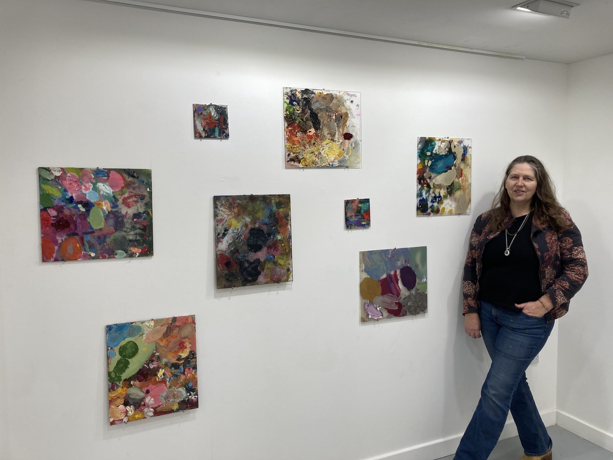 Woman in burgundy jacket and jeans standing beside white wall displaying eight colourful abstract paintings in various sizes.