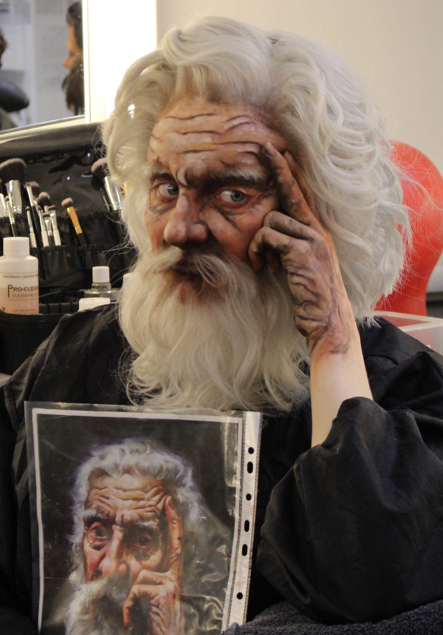 Person wearing make-up and prosthetics sits and holds a painting. The person has been designed to match the figure in the painting.