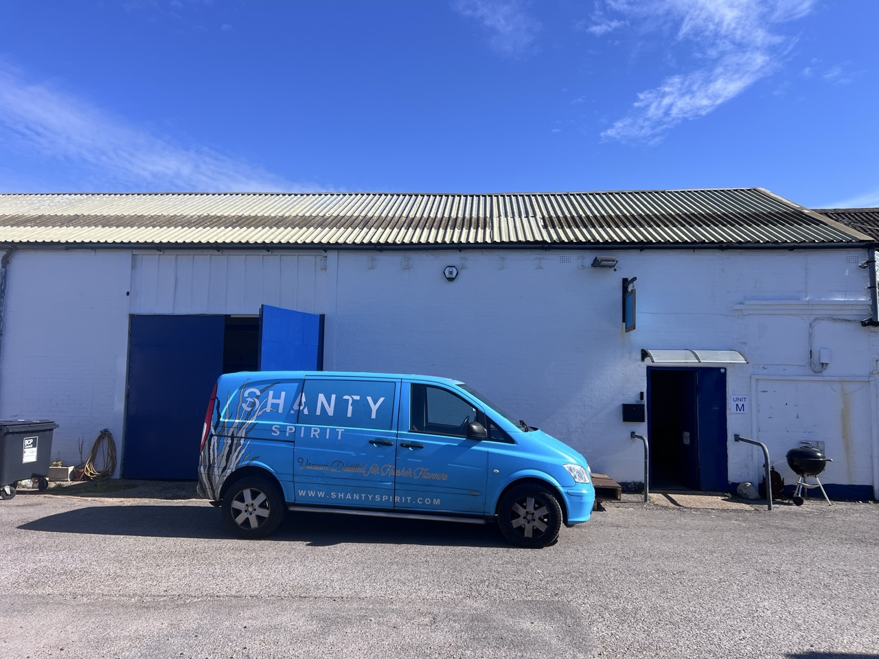 Blue van with Shanty Spirit logo outside a large white warehouse, blue sky above.