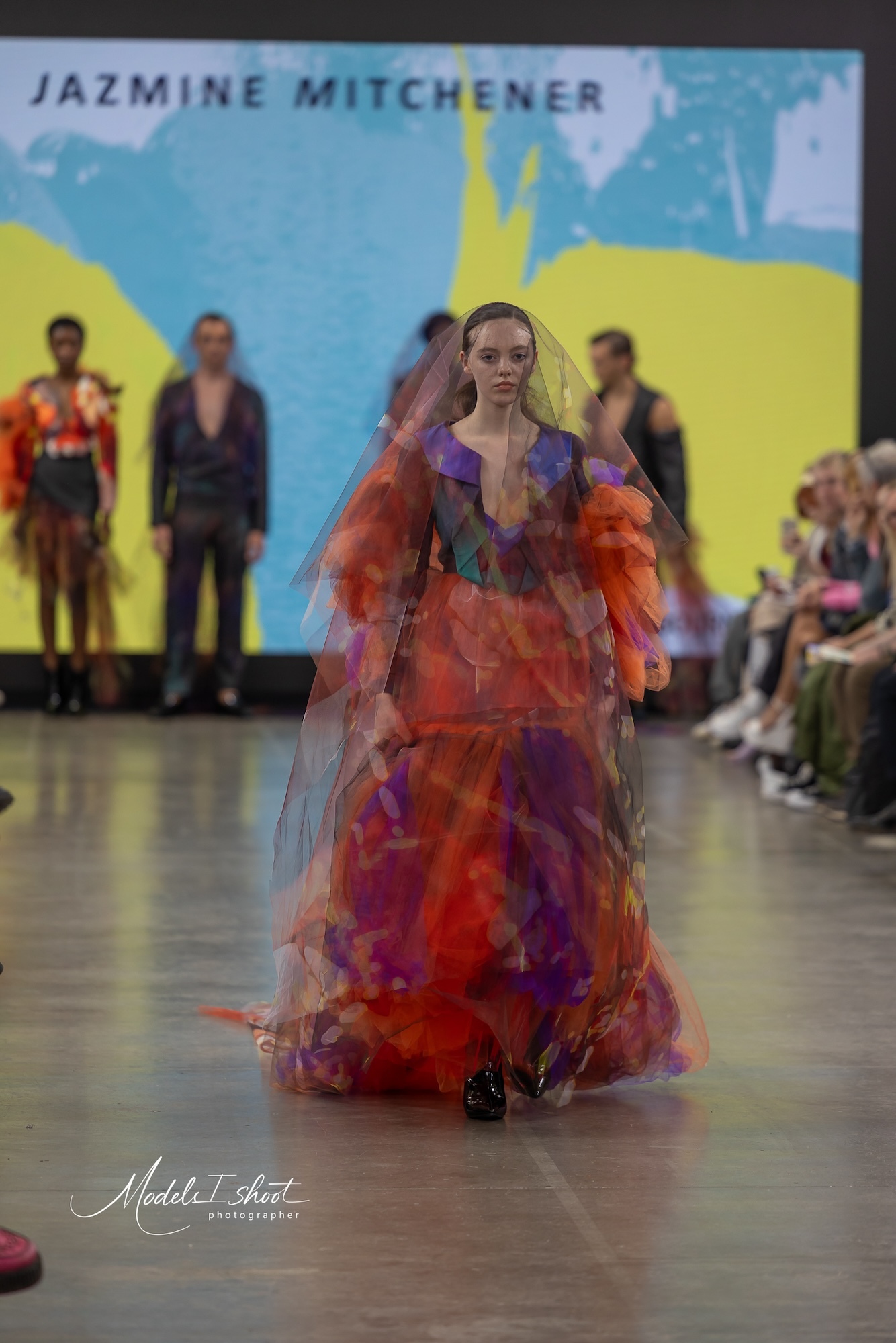 A model wearing a big red and purple outfit with a large veil walking down the catwalk at Graduate Fashion Week 2024.