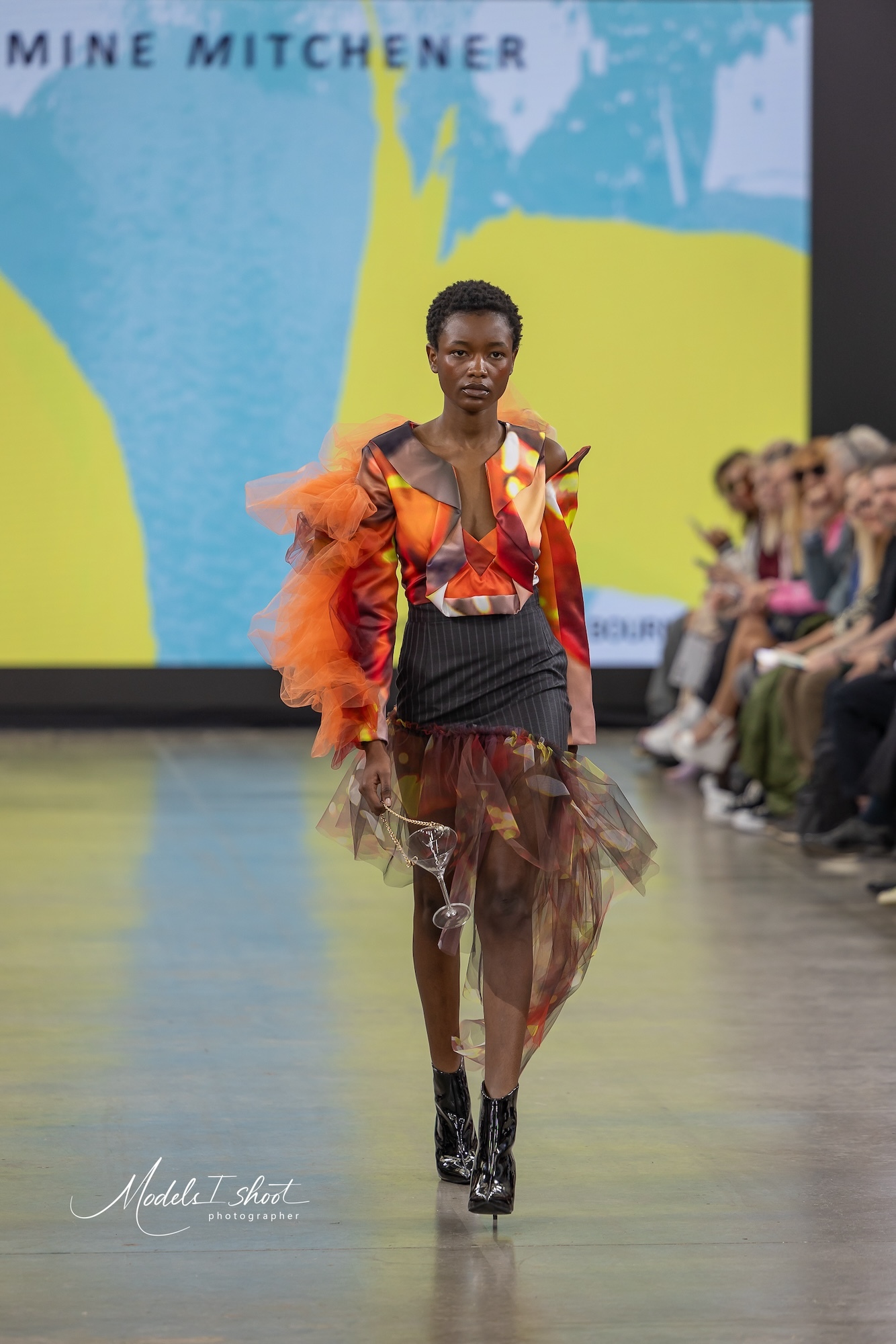 A model wearing a colourful abstract-patterned top and a pinstripe skirt holding a martini glass with a chain around it like a purse, walking down the catwalk at Graduate Fashion Week.