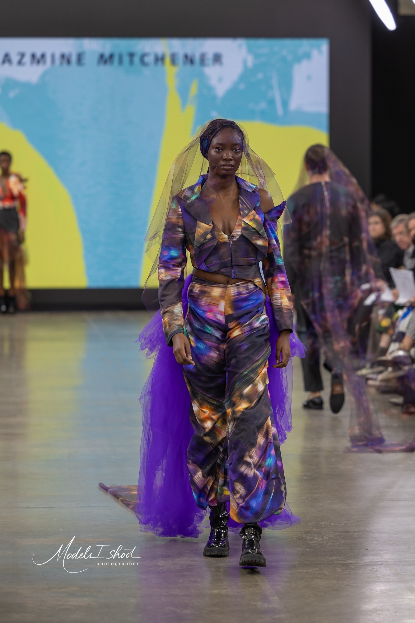 A model wearing an abstract-patterned outfit with purple skirt walking down a catwalk at Graduate Fashion Week.