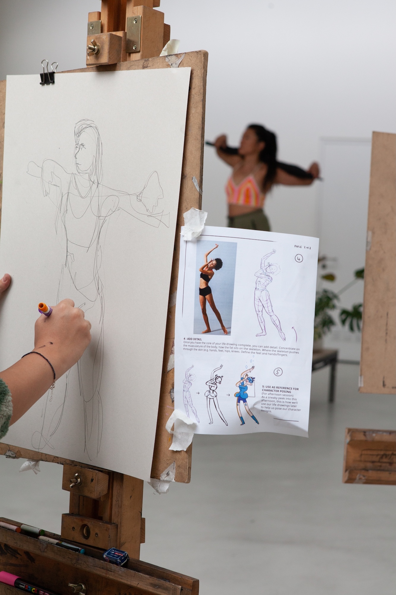 Wooden easel holds white paper with pencil figure drawing. Hand with bracelet sketching. Reference sheet with poses taped below. Blurred person in background.