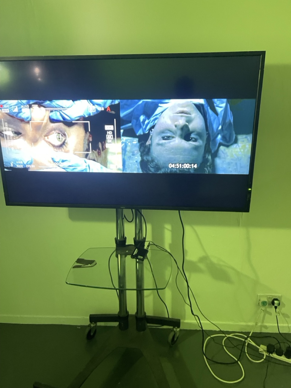 A flat-screen TV on a wheeled stand displays two close-up video images, with cables hanging below against a lime-green wall.