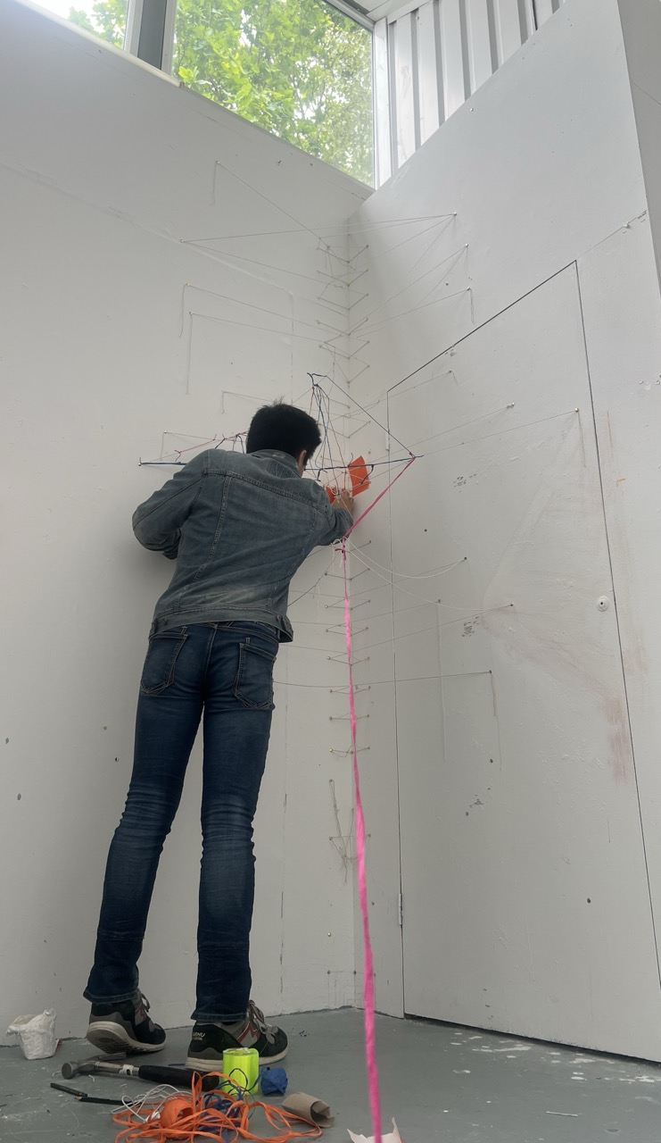 Seiji Suzuki interacting with abstract string art piece on a white wall, wearing a denim shirt and jeans.