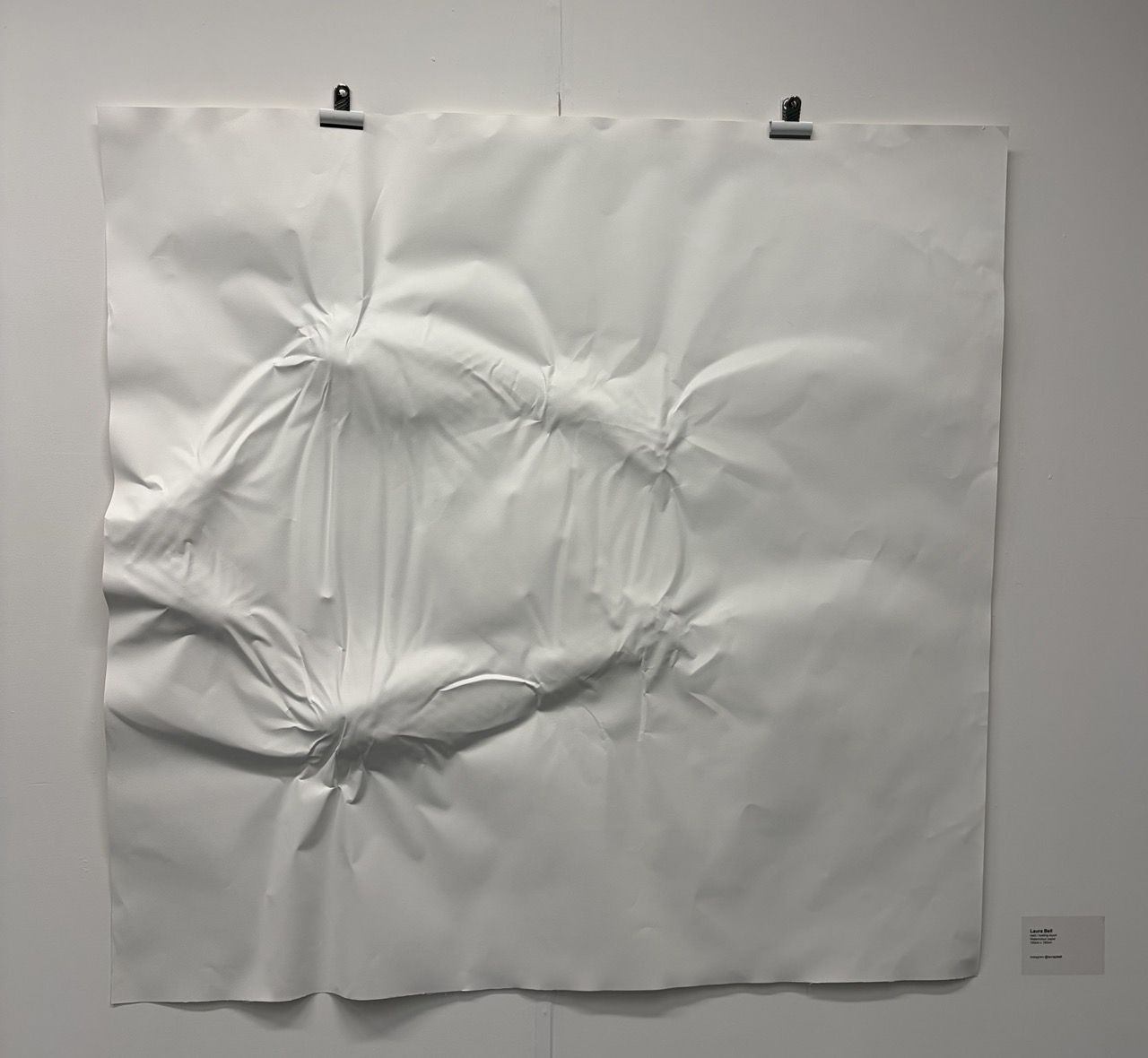 A large crumpled white paper sheet is pinned to a grey wall with two clips, with a small label mounted at the lower right.