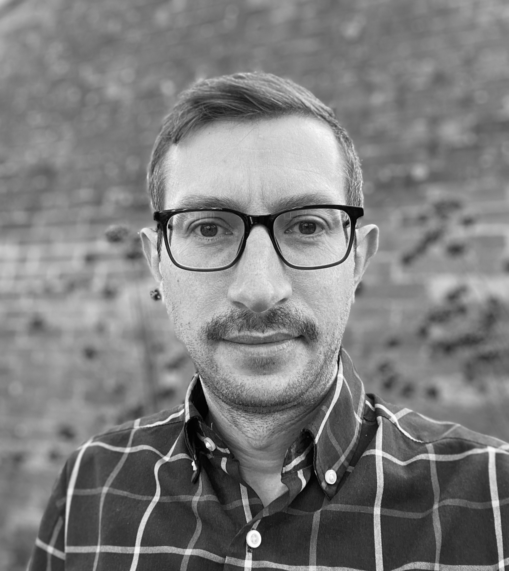 Black and white portrait of man with glasses and moustache wearing checked shirt against brick wall background.