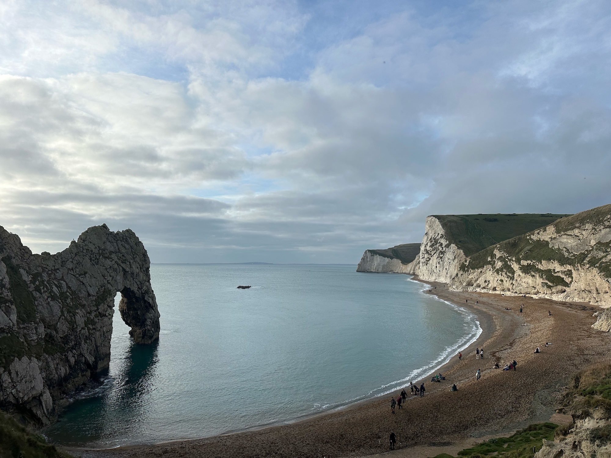 A panoramic view of a beach featuring the iconic Durdle Door rock formation on the left, with cliffs in the background. The calm blue sea meets a sandy beach populated with a few people walking along the shoreline.