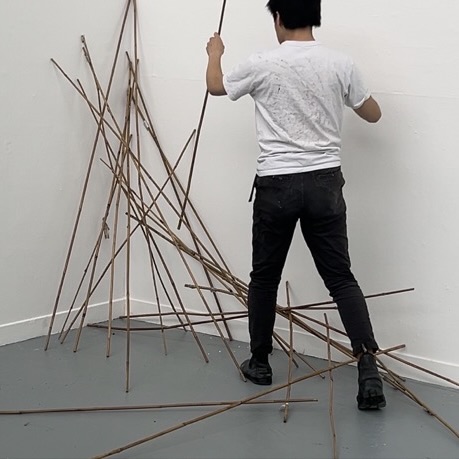 Seiji Suzuki stood wearing a white top and dark trousers, interacting with abstract bamboo stick art piece in a white room corner.