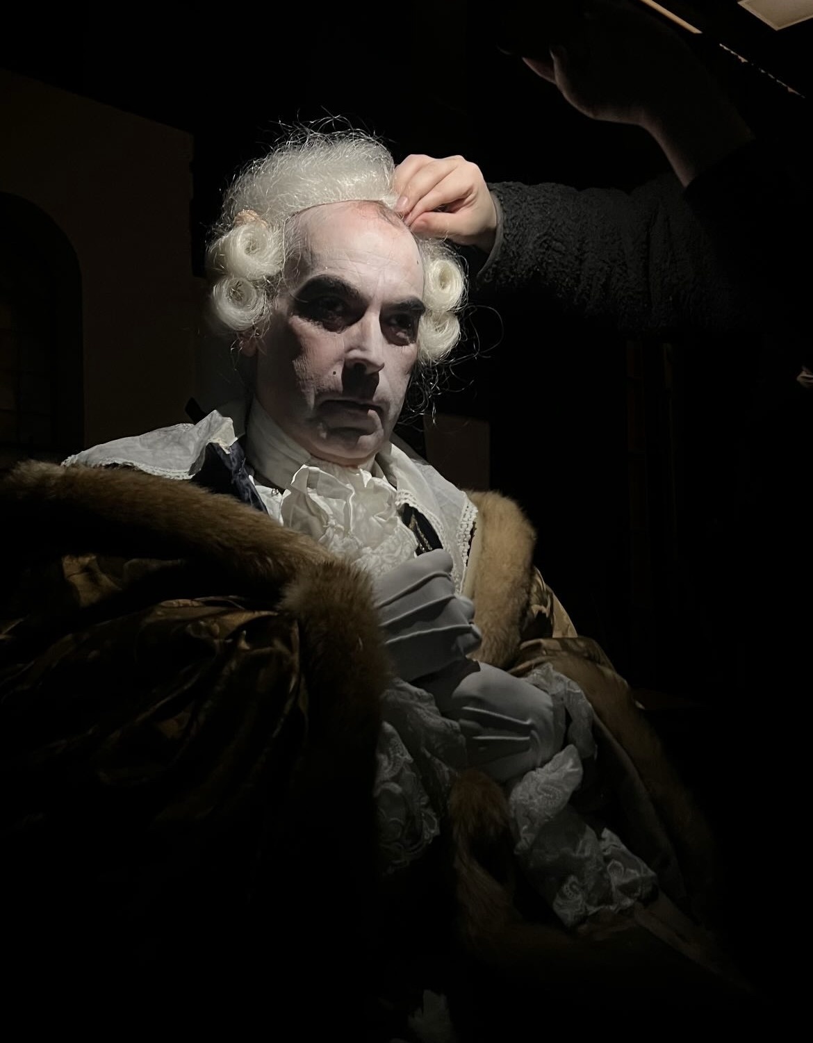 Close-up of a man being fitted with a 18th century-style powdered wig, under a spotlight.